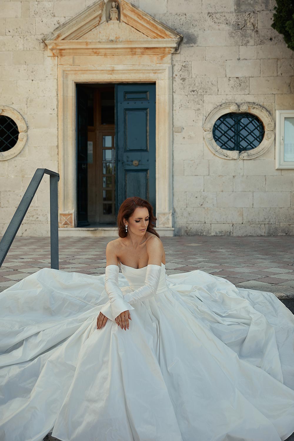 Marija, a layered dupion silk wedding gown of regal romance. Features a scoop neckline, draped bodice, detachable sleeves and bow, and a long train. From Vinka Design
